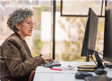 Older woman with curly gray hair and pink glasses working at a desktop computer in an office.