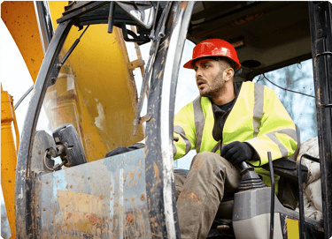 Construction worker in a red hard hat operating heavy machinery inside a cab.