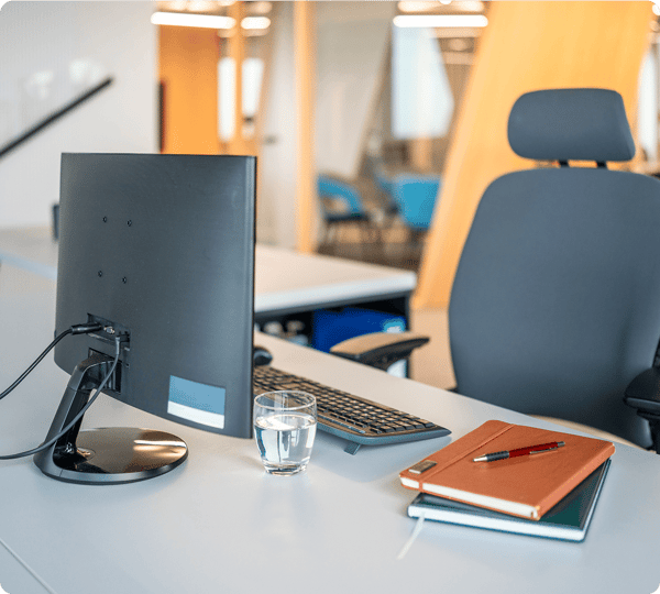 Modern office desk with a monitor, keyboard, glass of water, notebook, and pen in a bright workspace.