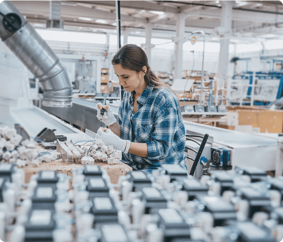 Woman in a factory assembling electronic components while wearing gloves and a plaid shirt.