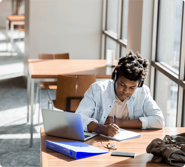 A student wearing headphones studies at a table with a laptop, notebook, and binder in a bright library.