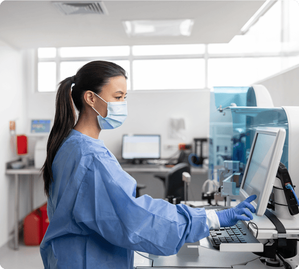 A healthcare worker in scrubs and a mask uses a computer in a laboratory setting.