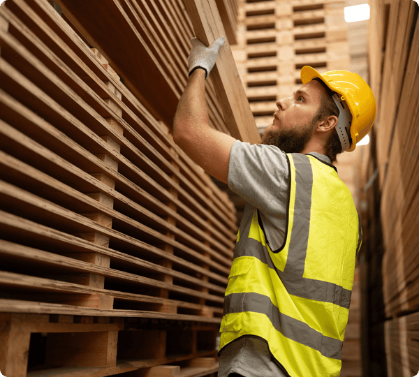 A worker in a safety vest and helmet inspects stacked wooden planks in a warehouse.