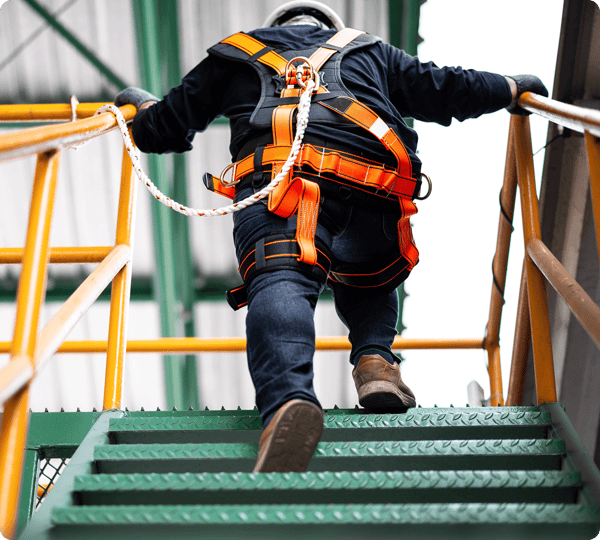 Worker in safety harness climbing green metal stairs on an industrial site, holding onto yellow handrails.