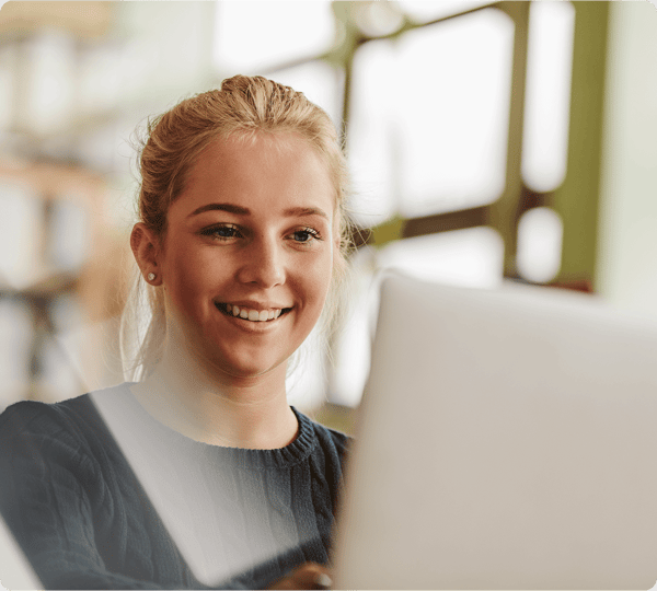 Smiling young woman using a laptop indoors with blurred shelves in the background.