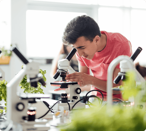 A student in a pink shirt examines a sample under a microscope in a science classroom with plants nearby.