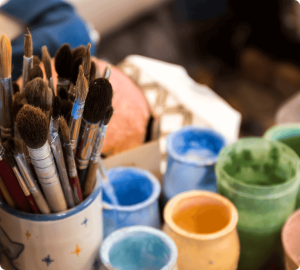 Assorted paintbrushes in a mug next to colorful ceramic cups and jars on a cluttered workspace.