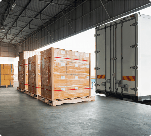 Several pallets of boxed goods wrapped in plastic near an open truck at a warehouse loading dock.