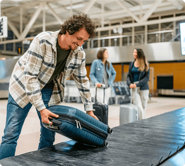 Man smiling as he picks up his suitcase from an airport baggage carousel; two women stand in the background.