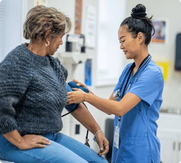 Nurse in blue scrubs checks the blood pressure of a patient sitting on an exam table.