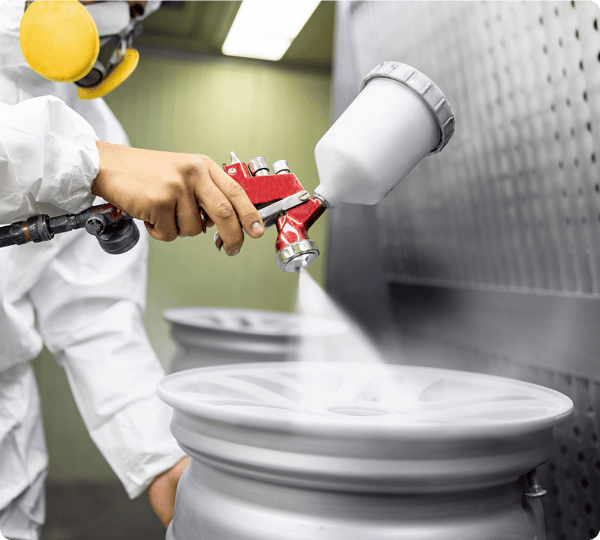 A person in protective gear spray-paints a car wheel rim with a spray gun in an industrial setting.