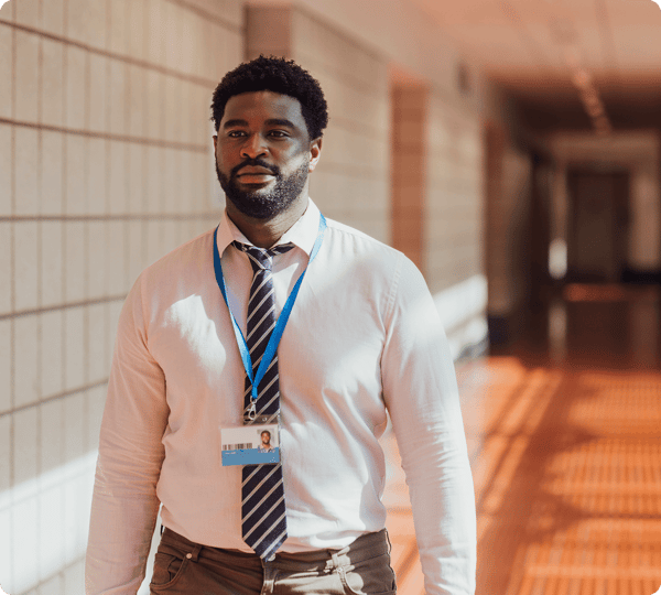 Man with a lanyard and ID badge walking in a sunlit hallway with tiled walls.