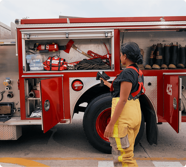 A firefighter stands beside an open fire truck, inspecting equipment stored inside its compartments.