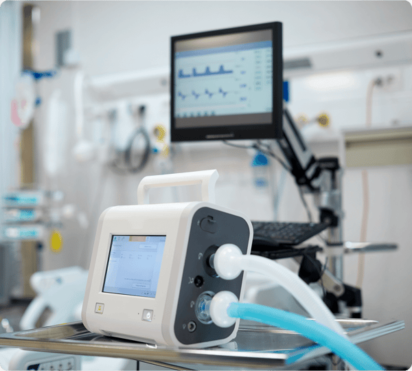 A medical ventilator with tubes is on a metal cart in a hospital room; a monitor displays patient data.