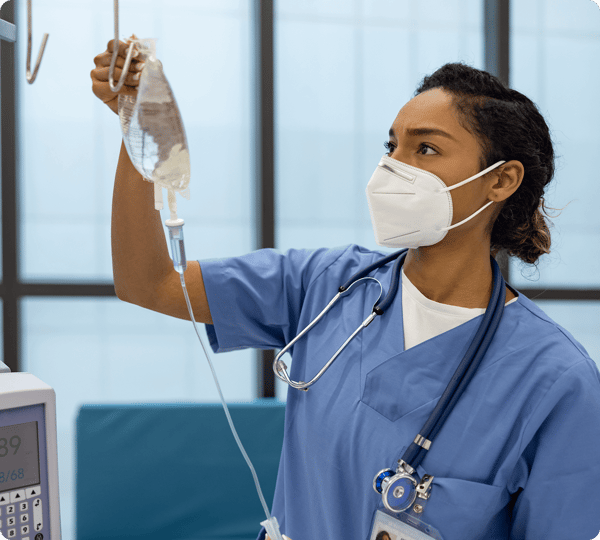 Nurse in scrubs and mask adjusting an IV drip in a hospital room.
