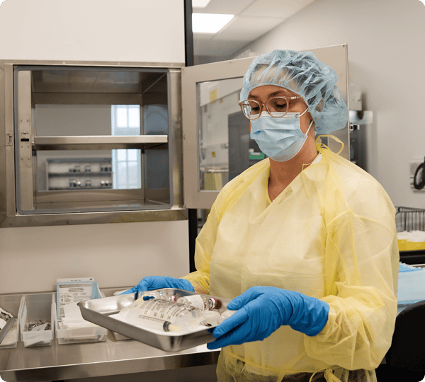 A healthcare worker in protective gear holds a tray with medical supplies in a clinical setting.