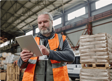 Man in safety vest using a tablet in a warehouse with stacked pallets and industrial equipment.