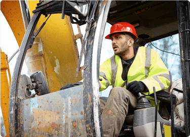 Man in a red hard hat and yellow vest operates construction machinery from the cabin.