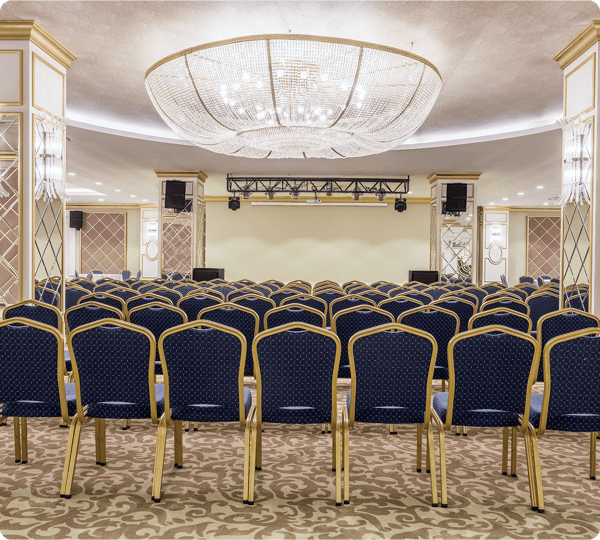 Rows of blue chairs face a stage in an elegant, empty conference room with a large chandelier.