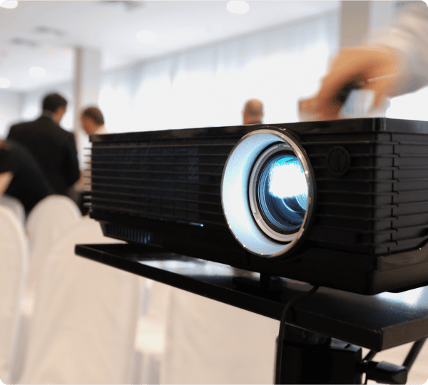 A close-up of a projector in a conference room, with people and white chairs in the background.