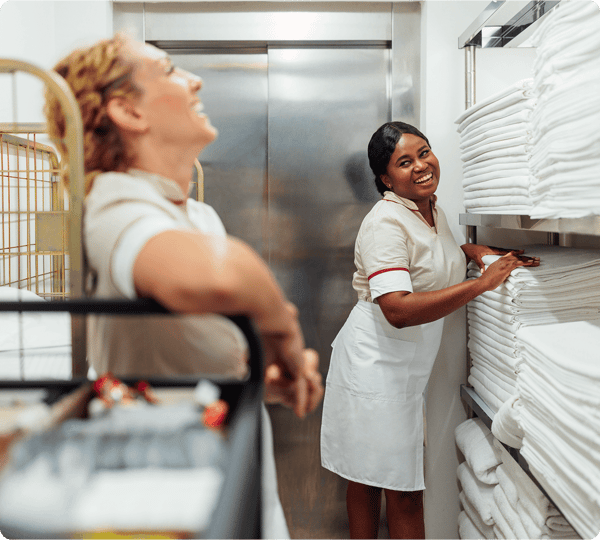 Two women in uniforms smiling and organizing white towels on shelves in a laundry or housekeeping area.