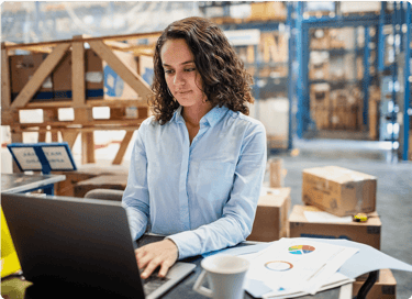 Woman working on a laptop in a warehouse, with documents and a coffee cup on the table.