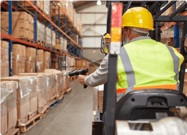 Worker in a yellow vest and hard hat scans boxes in a warehouse while seated on a forklift.