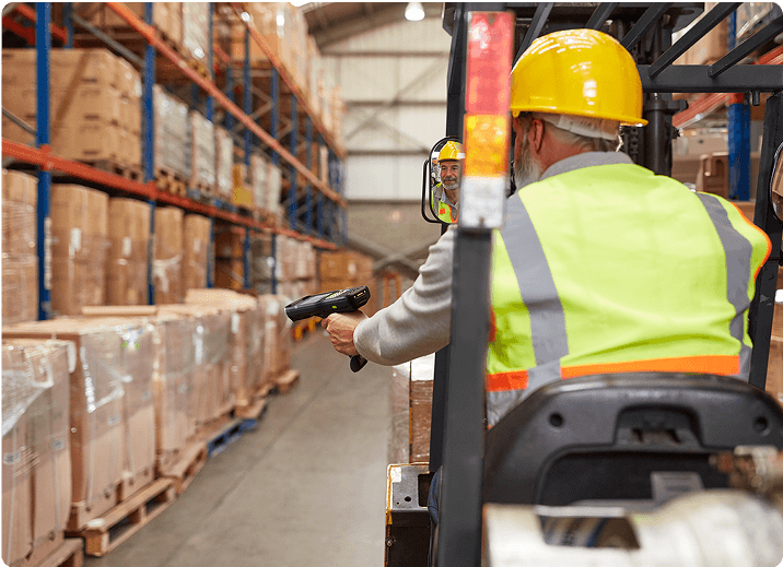 Worker in a yellow vest and hard hat scans boxes in a warehouse while seated on a forklift.