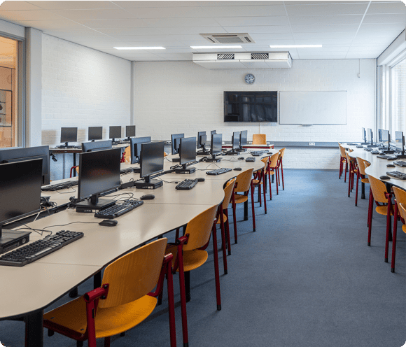 A modern computer lab with rows of desks, computers, chairs, a whiteboard, and a wall-mounted TV.