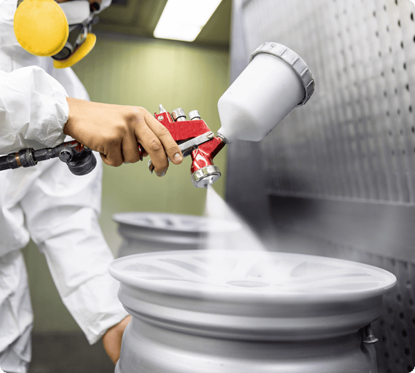 Person in protective gear spray paints a car wheel rim with a spray gun in an industrial setting.