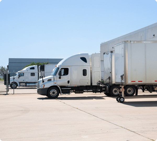 Two white semi-trucks parked on a concrete lot near a fence under a clear blue sky.