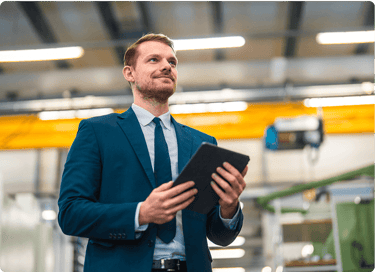 Man in a suit holding a tablet and standing in an industrial or factory setting.