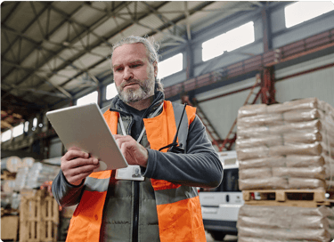 Warehouse worker in an orange safety vest uses a tablet in an industrial storage area with stacked pallets.