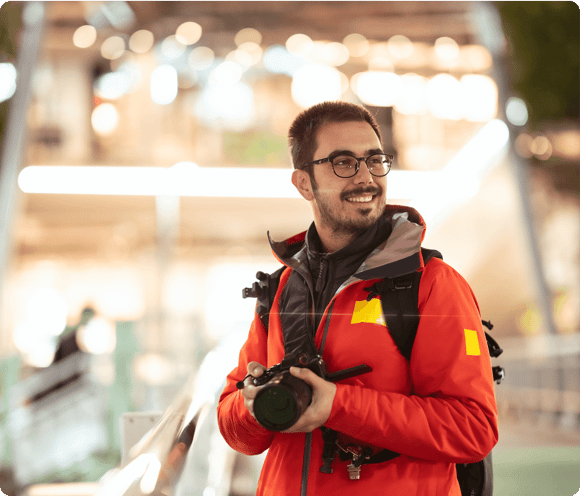 Man in a red jacket holding a camera and smiling, standing outdoors with blurred lights in the background.