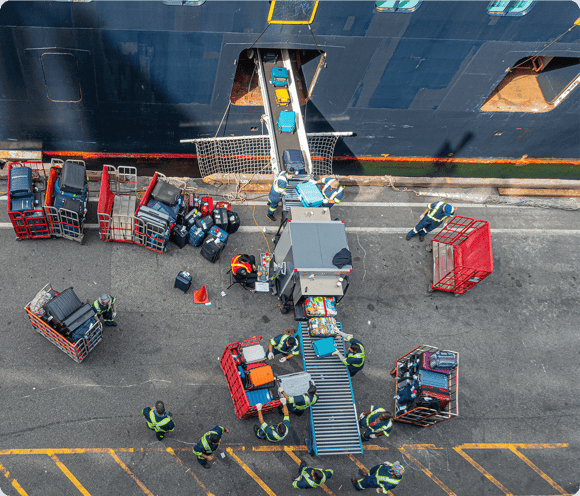 Aerial view of workers loading luggage onto a ship using conveyors and red luggage carts.