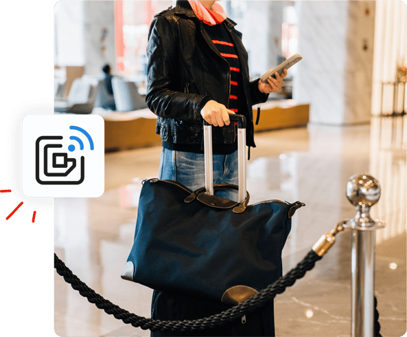 A person in a hotel lobby holds luggage and a phone, standing near a rope barrier with a digital icon shown.