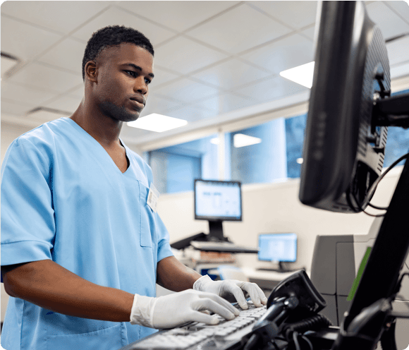 A healthcare worker in scrubs and gloves types on a computer in a medical office.