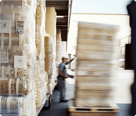 A worker moves a wrapped pallet of goods off a truck loaded with similar packaged items in a warehouse area.