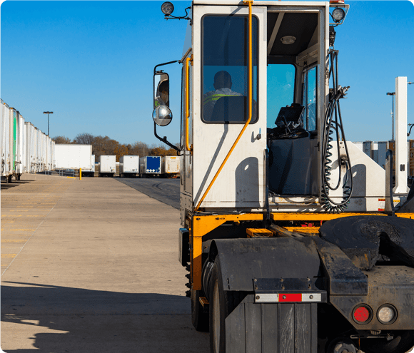 A truck driver sits in a cab, preparing to move trailers in a sunny industrial lot with many trucks.